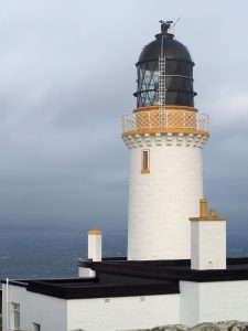 A tall white lighthouse against a grey sky. The tower is white and cylindrical. On top is a brown-painted balcony, and above that is a black dome which, presumably, holds the light. 