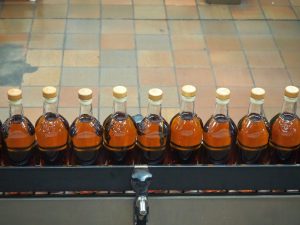 a row of bottles on a conveyer belt. Each holds a golden-brown liquid and has a lid.