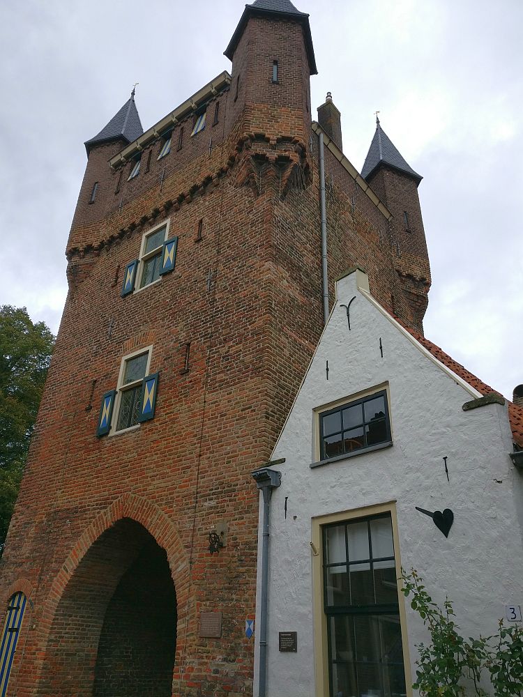 The city gate is a tower of red brick, with a small turret on each corner at the top. The small house next to it is simple and white with a peaked roof and two windows. 