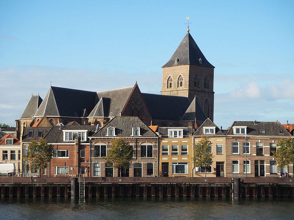 A row of old, square, three-story houses lines the quay with, behind, them, a large, clearly medieval-era church with a blockish square tower that has a pointed roof.