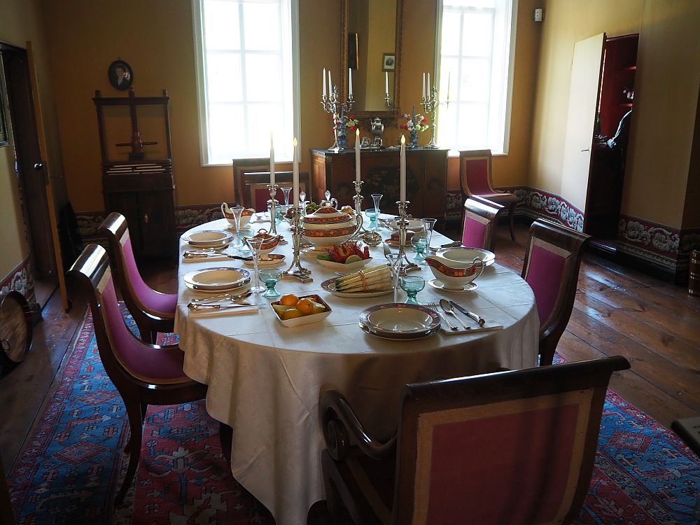 The Menkemaborg dining room has a relatively modest looking table, set for six people, with a white tablecloth, silver candlesticks, and what looks like fine china and silverware. The chairs are simple yet elegant, with gently curved backs and upholstery in dark reddish-purple. The room's floor is wood boards, but the table and chairs sit on an oriental carpet.