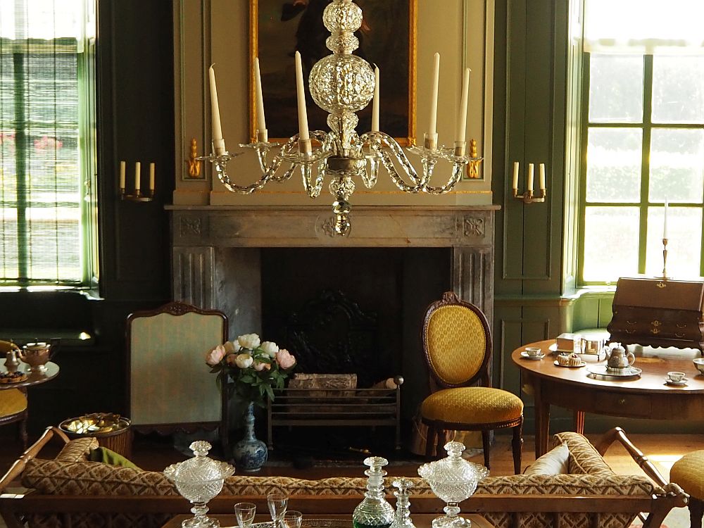 A pleasant light room in Menkemaborg, done in browns and light greens and yellows. This view shows the back of the sofa looking toward the fireplace. The painting above the mantel is obscured by the simple but elegant crystal chandelier that holds candles. A yellow chair near the fireplace, next to a table set for tea. A bouquet of flowers, also next to the fireplace on the floor in a porcelain vase. 