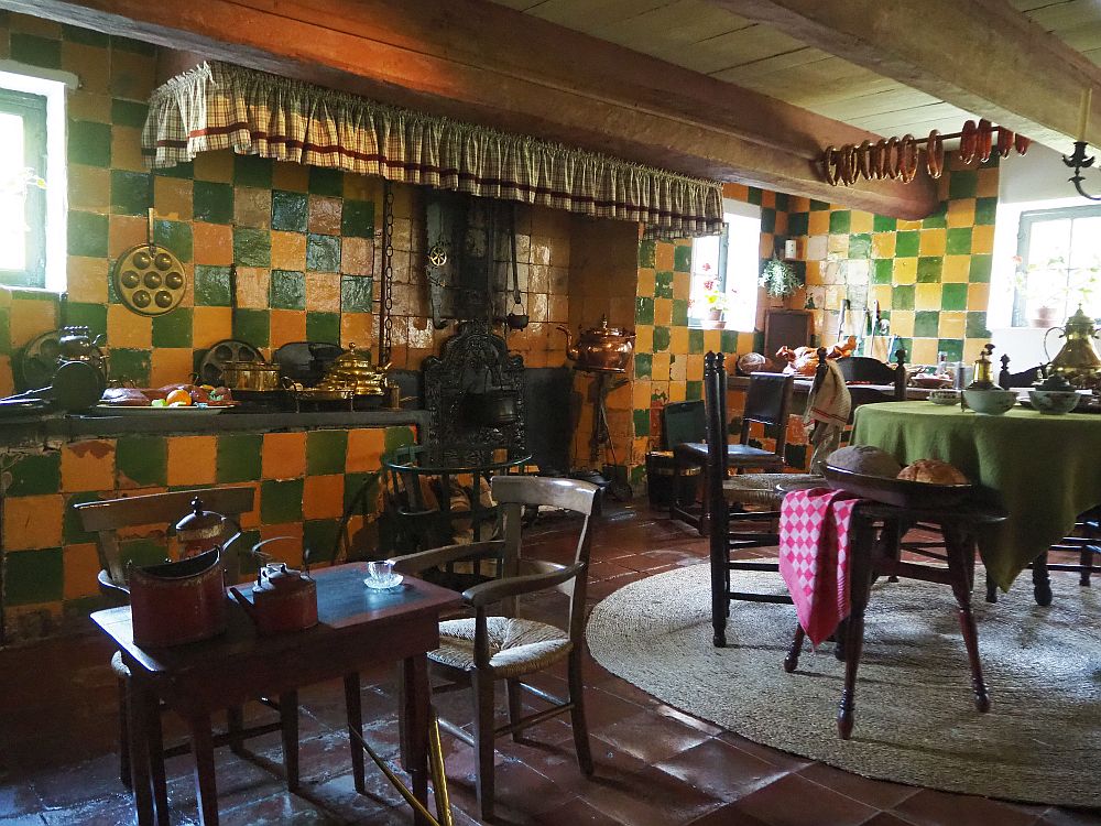 the kitchen's walls are tiled in green and yellow, while the floor is simple dark red tile. In this picture are two tables: on the left is a chld-sized wooden table with two chairs and on the right a regular table with a green tablecloth. Behind is a counter with copper pots and other kitchen items next to a fireplace with an iron stove in it and a door in the wall, presumably for baking. A sort of ruffled curtain hangs around the chimney above. The ceiling is quite low and has heavy beams across it. Sausages hang from a rod between two of the beams. 