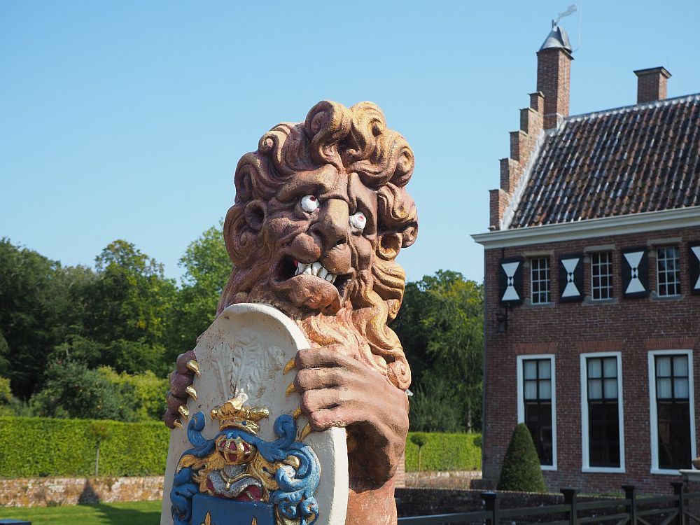 One of the lions at Menkemaborg: he stands upright, clutching a coat of arms in his front claws. He looks to the right, with his eyes looking upward. the lion is painted brown except for the eyes and teeth, which are painted white, and gold claws. The coat of arms is white, with details in gold and blue.