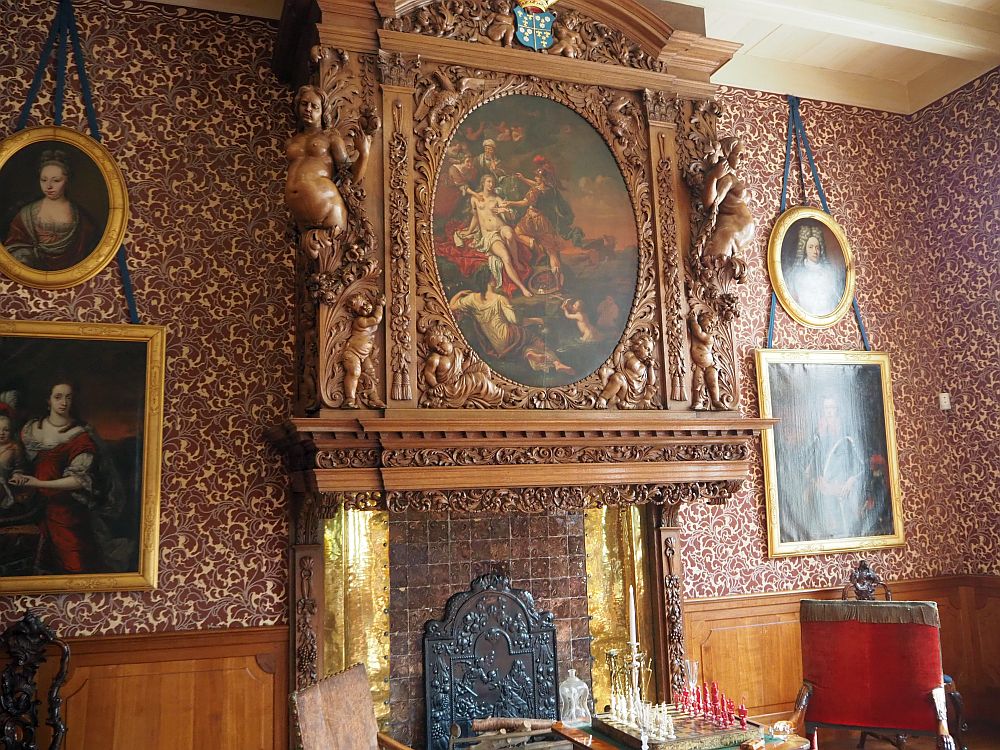 From mantel height to the ceiling, the mantelpiece is carved, with a frilly rim below, naked female figures on the corners, and an ornately-carved oval frame in the middle, framing a scene, probably from classical mythology, invloving a naket woman surrounded by three dressed people, with another naked woman and a baby reclining at her feet.