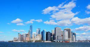 bright blue sky and fluffy clouds above a view of the NYC skyline as seen from the south. The new World Trade center building is visible along with lots of other skyscrapers.