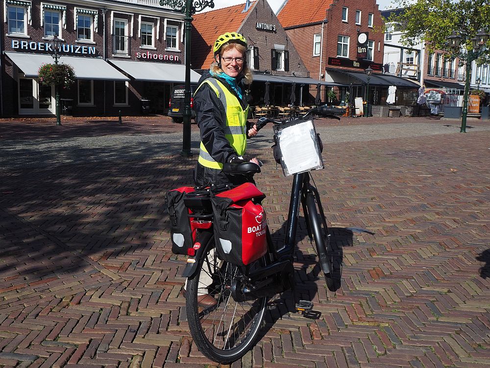 Sandra, tour leader on our Boat Bike tour, stands holding her bike, which has red panniers on either side of the back wheel. She is wearing a bright yellow safety vest and a raincoat under it, and her helmet is also bright yellow. She wears glasses and has blond hair. She smiles at the camera. 