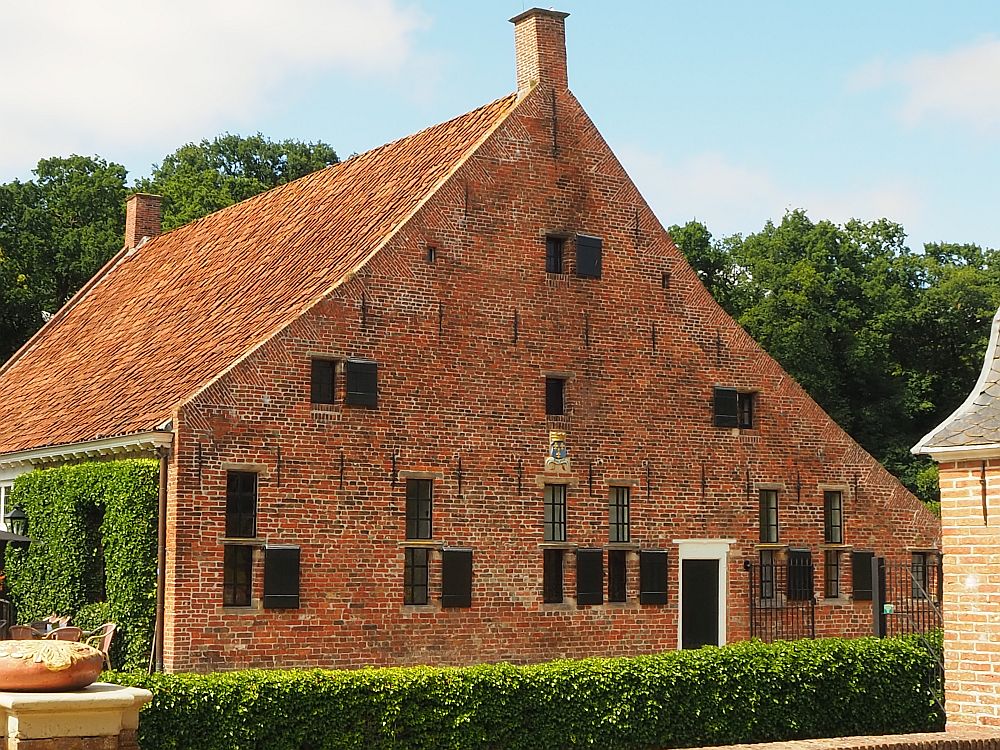 The photo of Het Schathuis at Menkemaborg in Uithuizen shows the side of the building: a triangular shape with a flat redbrick face. Small windows pierce it here and there. A small part of the front of the building is visible, covered in ivy, and just on the edge of the picture one of the chairs of the outdoor part of the cafe is visible.