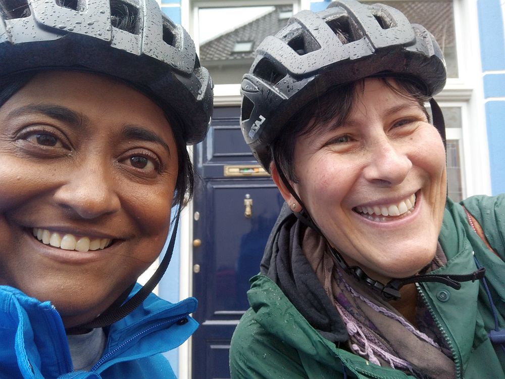 Shobha on the left, looking at the camera, me on the right, looking to the side. Both smiling, wearing black bike helmets and raincoats, which are wet.