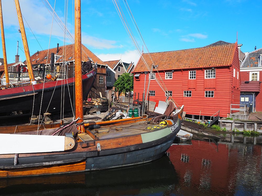 In the foreground a wooden botter lies at anchor. Behind is a bright red building with simple square windows.