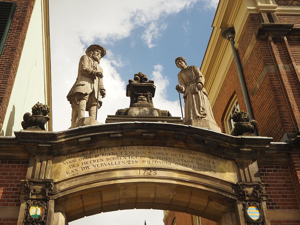 The arch has text carved into it and the date 1723. On top of the arch stand two statues: on the left, a man in jacket and hat, leaning on a cane. On the right, a woman in a long dress, also leaning on a cane.