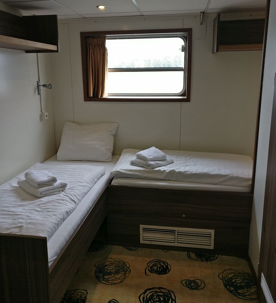 Two twin beds, in our cabin on our Boat Bike Tour, bedding folded on top. A window on the back wall.