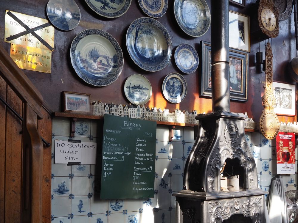 The upper half of the wall is brown wood panelling hung iwth china plates. The bottom half is tiled with delft tiles, blue figures on white. A narrow shelf divides the two halves and holds a row of delft ceramic houses. In front of the wall is a cast-iron stove with a stovepipe extending straight upward. This cafe was the first stop on the Jordaan food tour.