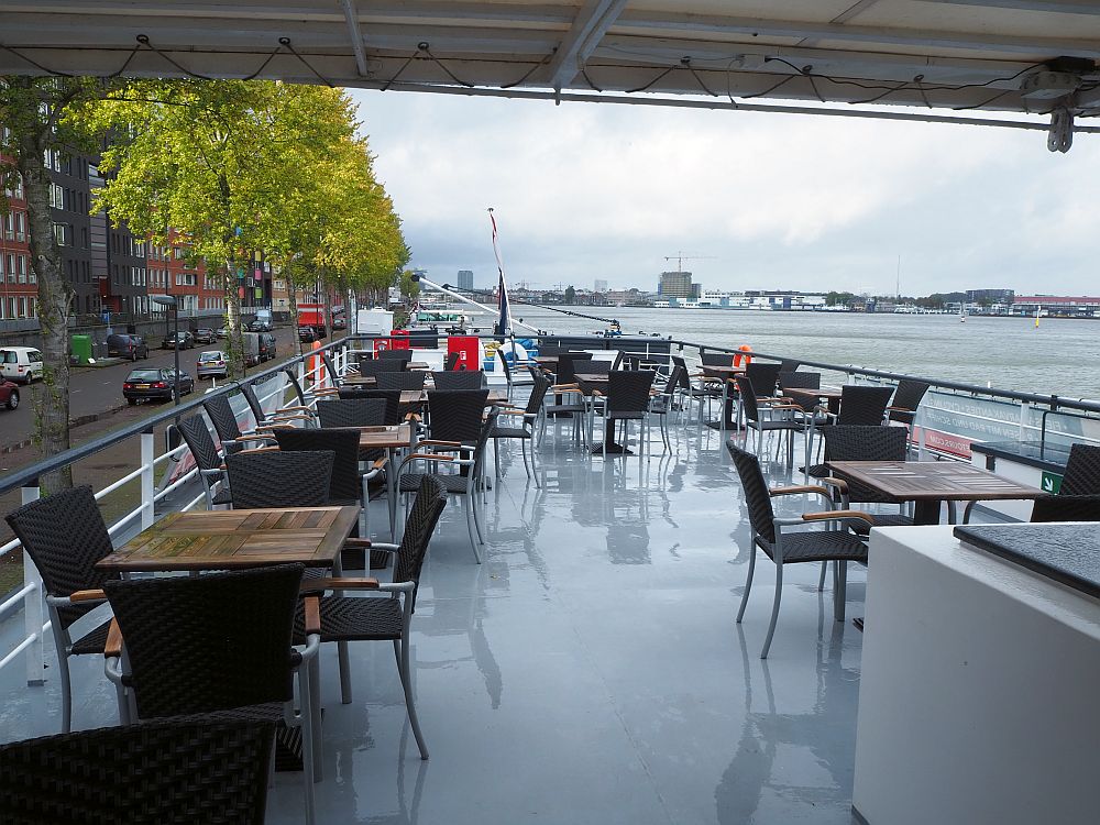 A flat, grey deck with railings around the edge. Wooden tables with black chairs around them.