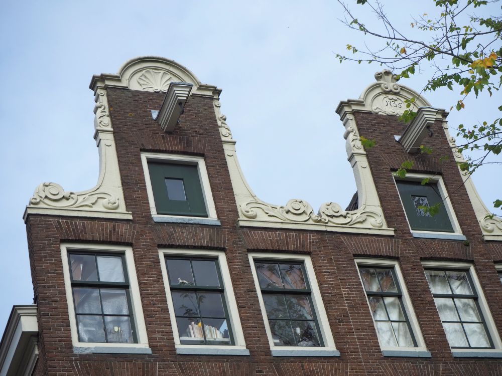 These two gables are vertical red-brick rectangles, but each has a white decorative element surrounding it, with curls at the base, a pillar-like shape on either side, and an arch above the window. The arch on the left gable has a shell shape. The arch on the right has the date: 1735. Each gable has a beam sticking out with a hook on it.