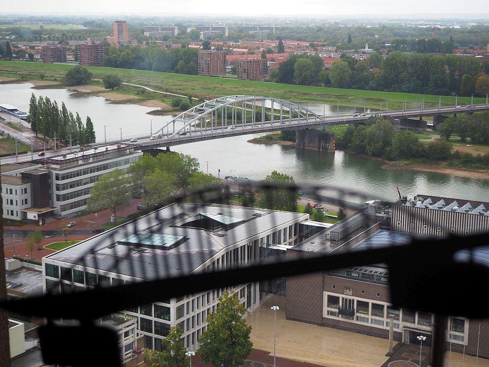 The river crosses the picture, and the arched bridge crosses the river. In the foreground is a blurry outline of the bridge -- a decal on the tower window. Below on the near bank of the river are recent buildings, as well as in the distance on the far side of the river.