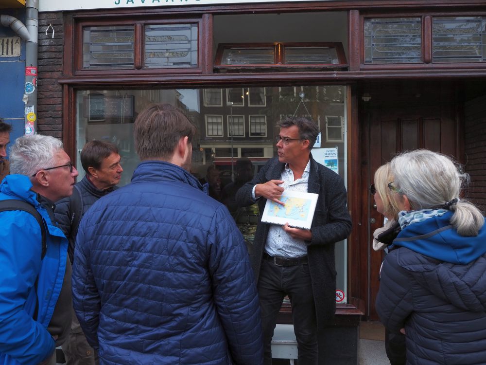 A man (Kenny) in a white shirt and dark pants and jacket stands facing the camera, holding a small world map and pointing to it. A group of other people stand in a half-circle around him.