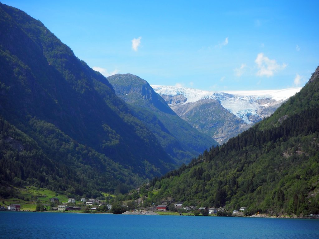 A view in Norway: blue water in the foreground, a small cluster of houses on the shore. On either side of the village, huge tree-covered mountains. In the background, even higher mountains, covered in snow.