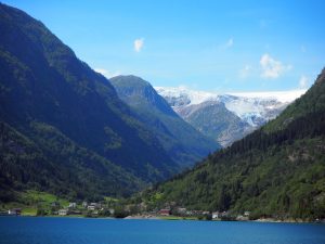 blue water at the bottom of the picture, dark green hills on either side of a valley rising from the water. More hills behind, and the furthest mountain is snow-topped.