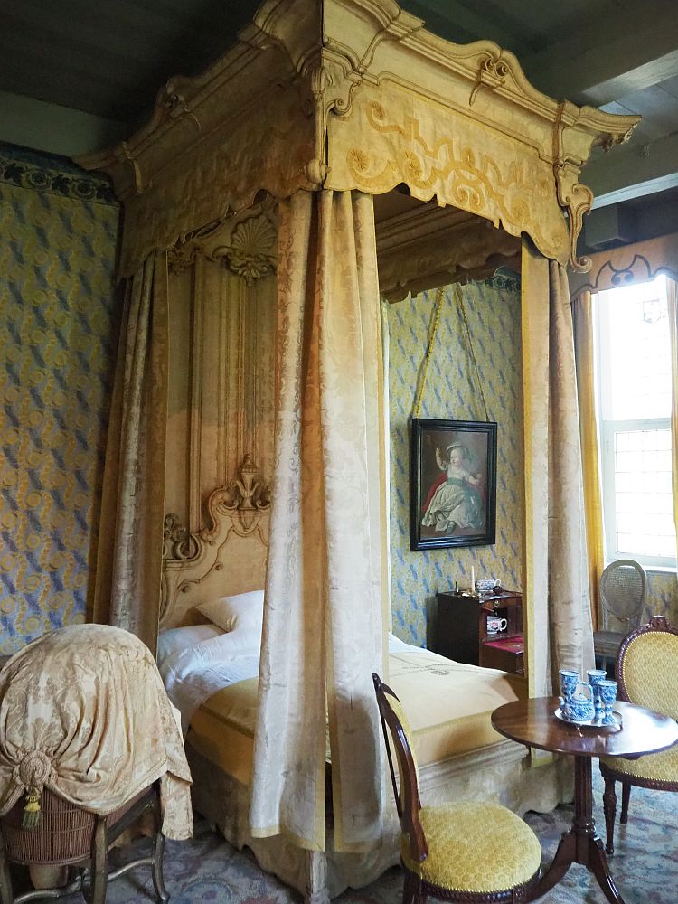 This photo is vertical of necessity because the roof of the four-poster bed reaches the high ceiling. The woodwork at the top as well as the curtains at each corner are pastel yellow. The headboard of the bed is partly visible and is ornately carved, also painted yellow. The wall behind had yellow wallpaper (or silk?) with a pattern of blue on it. Next to the bed is a small cupboard and a painting of a child hangs above it. At the foot of the bed is a simple round table with two upholstered yellow chairs and a small tea set in its middle. 