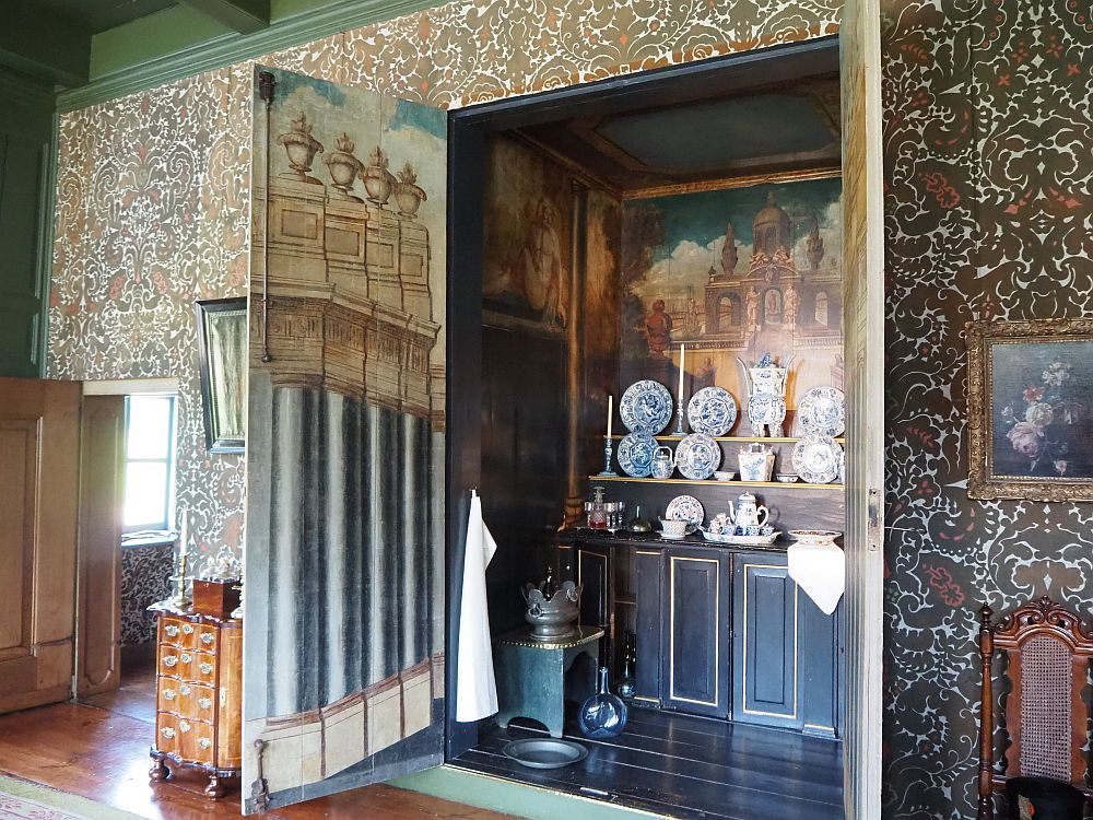 The doors to the buffet (more like a big closet) open outwards. Inside is a sideboard, with cabinets below and shelves above, covered with a blue and white porcelein set. The walls of the inside of the closet and the insdes of the door are painted with classical scenes: some sort of arched building on the back wall and large columns on the door.