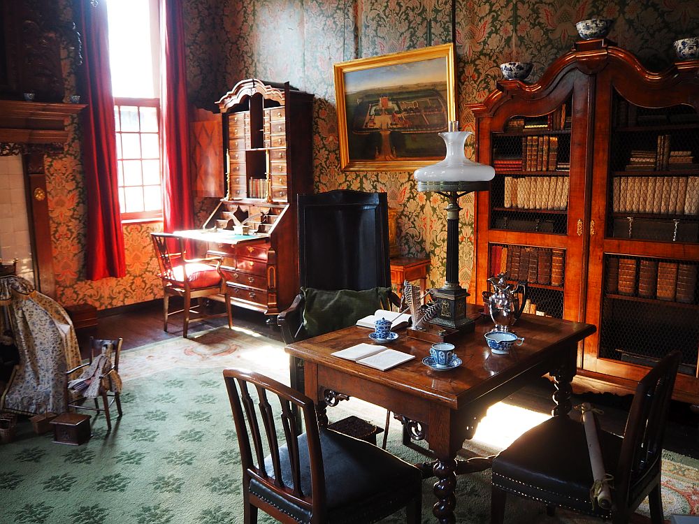 In the foreground, a square table with two chairs. On the table a lamp and teacups. Behind it, against the wall on the right, is a wooden cabinet with glass doors. On top are some pieces of blue and white porcelain. Inside are rows of books. IN the background is a secretary, open for writing. It has many drawers above and also slots for papers. A simple cushioned chair sits in front of it. Between the book cupboard and the secretary hangs a painting, though it is unclear what it portrays. ON the left, on the edge of the photo, is a small doll-sized chair holding an old-fashioned doll. 