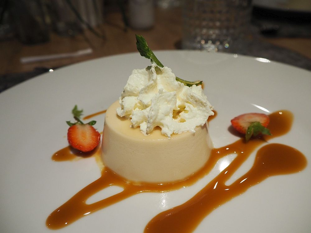 The panna cotta is perfectly formed from a round mold. It has a bit of whipped cream on top, two halves of a strawberry beside it, and a dribble of caramel sauce.