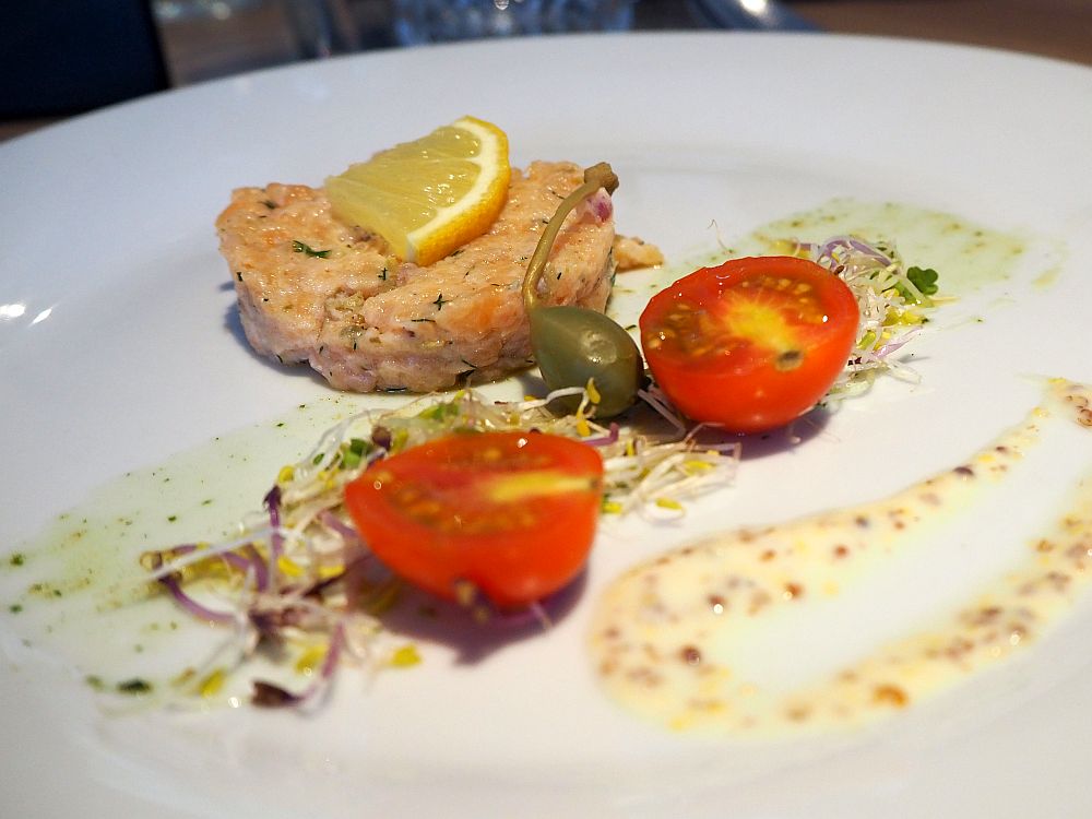 A dollop of salmon with a slice of lemon on top. Next to it, two halves of a cherry tomato, a fresh olive, and some sprouts. All of it is dribbled with dressing. 