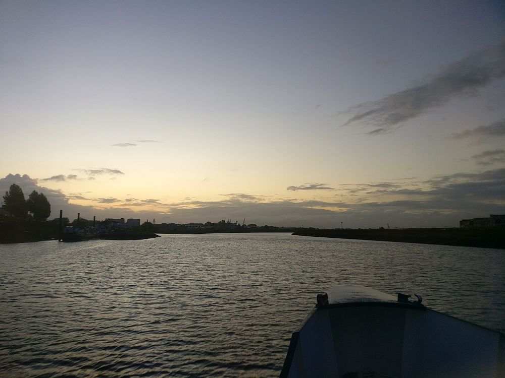 The bow of the boat for our boat bike tour in the foreground, in silhouette. Water ahead, with flat riverbanks in the distance, also in silhouette. The sky and clouds are lit up before the sunrise.