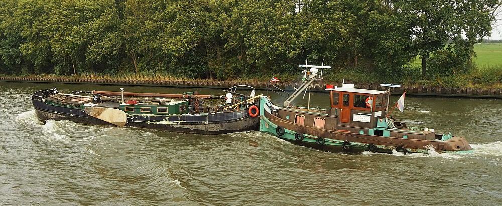 An old-fashioned little tugboat pushes another boat, which is a rundown-looking older wooden Dutch boat, the kind with sideboards. The mast is lying on top of it.