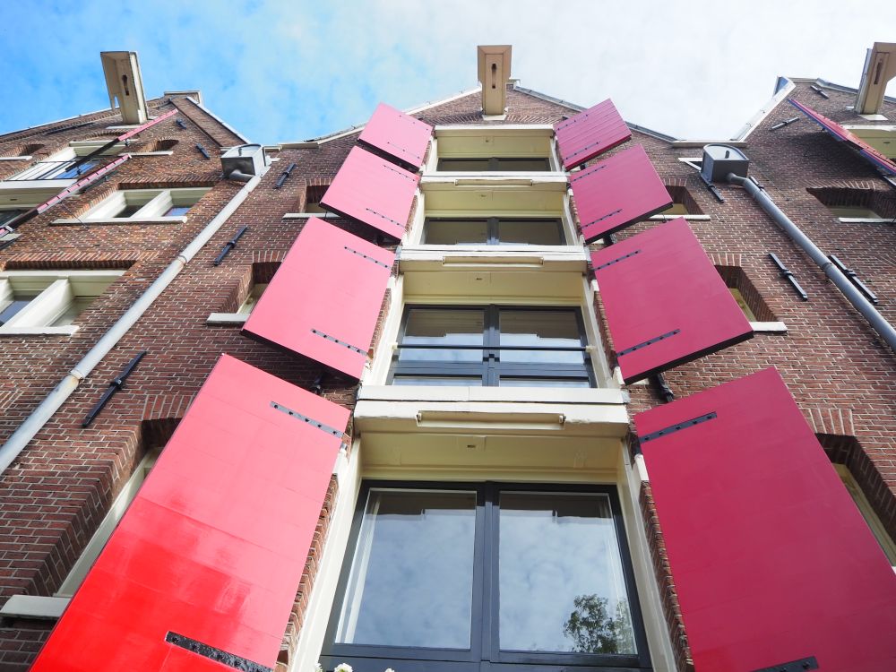 Looking up at the building, the center of the picture is a vertical column of windows, each with open red shutters on each side. 