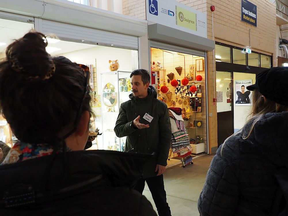 Archie, facing the camera, stands in front of a row of shops inside the market. In the foreground are the backs of a few peoples heads. He is telling us about tradtional Latvian food.