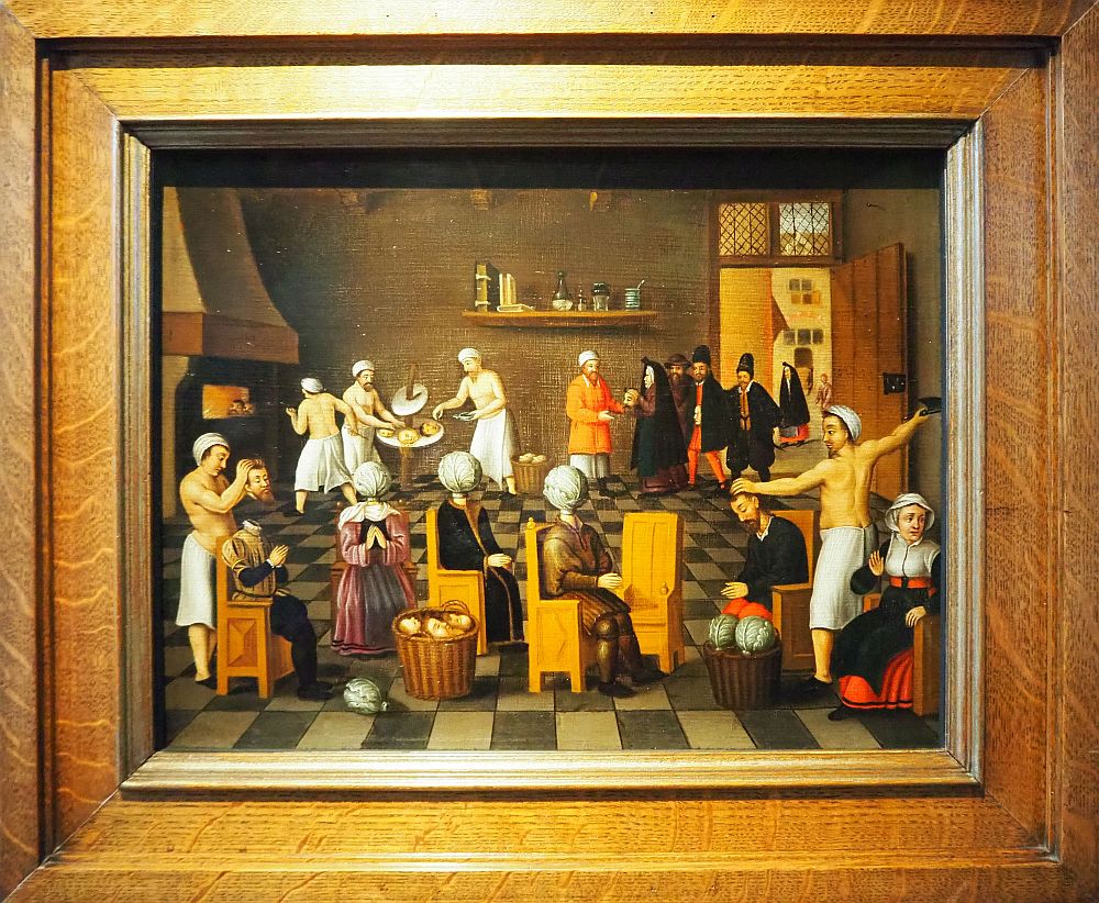 In front are the people with cabbages rather than heads. They sit in a row of chairs. A man on the right is holding a knife high, about to slice off a man's head. A man on the left is holding a head over a sitting torso, about to place the head back on. Behind on the left is a fireplace and a shirtless man is placing heads inside it with a long paddle. Two other men are preparing heads on a table. On the right rear people in black coats are lined up and a man in a red coat receives them.