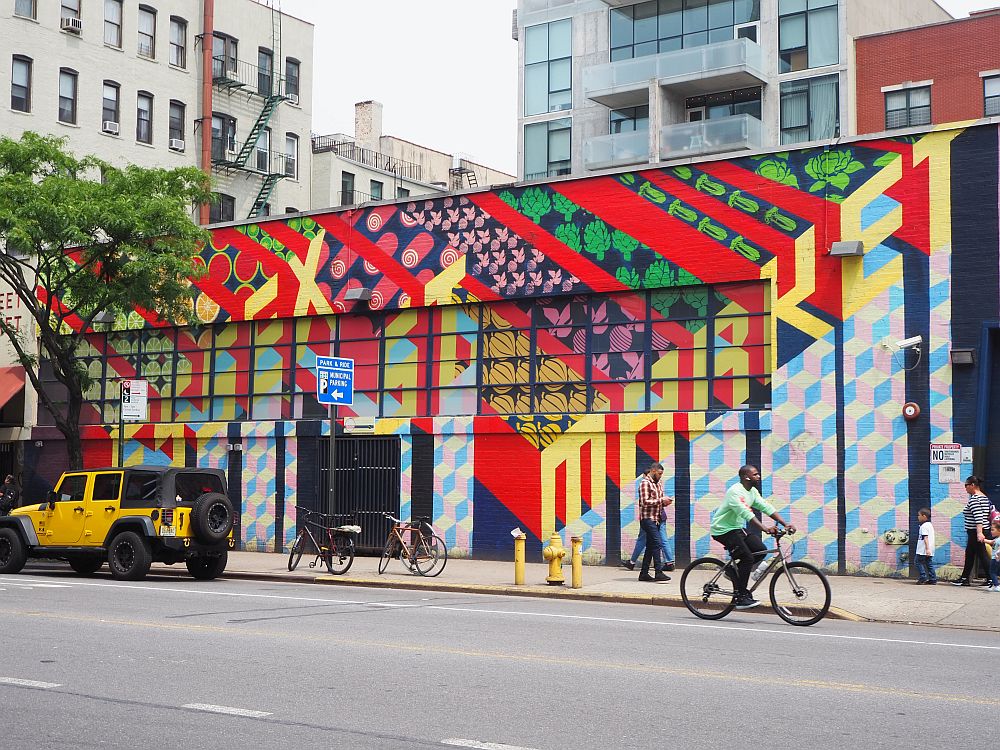 Taken from across the street, this photo shows a one-story building with a few taller buildings visible behind them. The Essex Street market, a stop on one of the Tenement Museum tours, is painted in bright colors, with broad stripes of red and, between the stripes, repeated images of items that are sold in the market, e.g. artichokes and pickles. Across the strips the words Essex Street Market are visible painted in yellow in a slanted line.