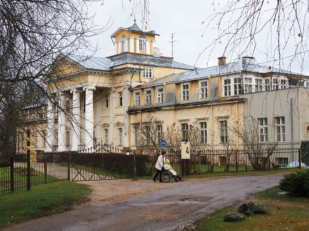 The mansion is light yellow and has classical columns in front of the main entrance, holding up a triangular pediment. The center part is 4 stories tall with a small square structure on the roof. The two wings are lower, just two stories. A woman pushes another woman in a wheelchair in front of the manor.