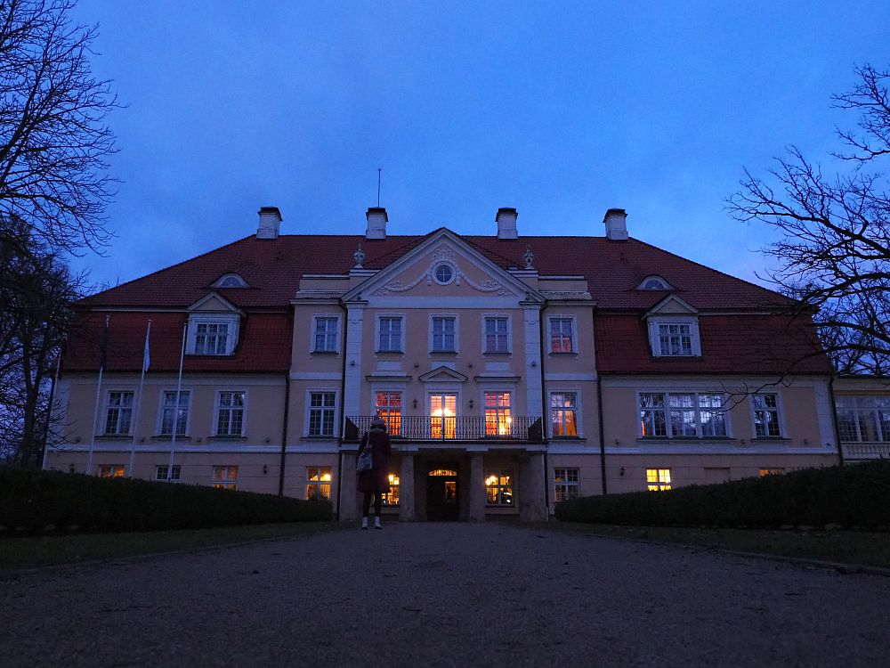 The symmetrical manor house is a pastel orange color with white edging and a reddish roof. It is four stories tall with a grand, decorative entrance. The sky is dark and lights blaze from the windows on the first two floors.