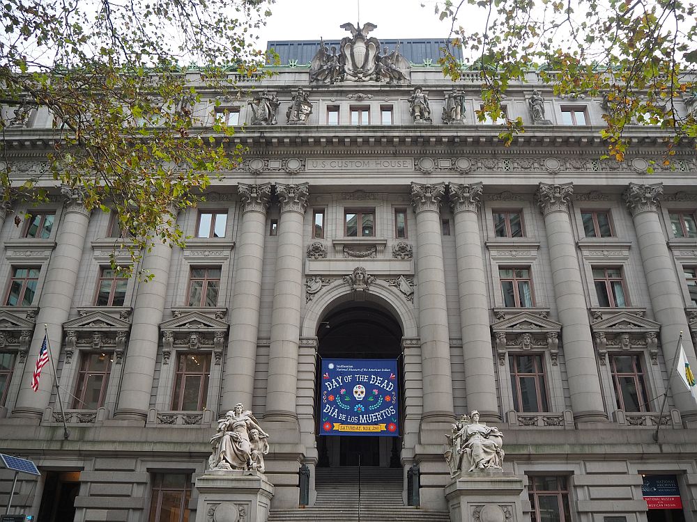 A very grand front to the building: granite, with corinthian columns and ornate sculptures on either side of the entrance stairway. A banner hangs above the entranceway reading "Day of the Dead. Dia de los muertos" 