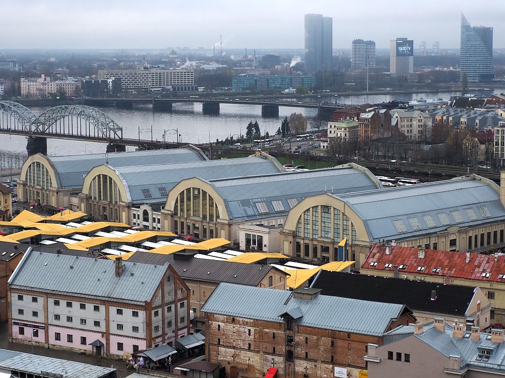 View from a high building shows the 4 half-cylindrical buildings in a row. In front are some rectangular brick buildins a few stories tall. Behind is the river and three bridges crossing it are visible. Low-rise buildings behind the market on this side of the river, a few high-rise buildings on the other side. 