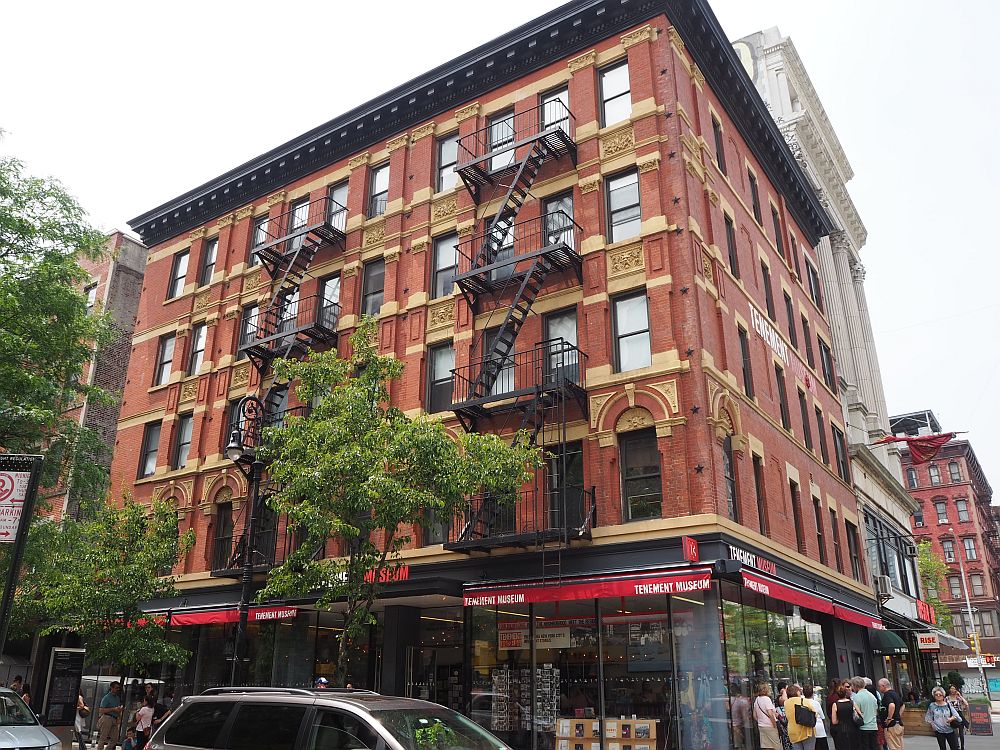 A corner building, red brick with decorative elements between and above the windows in white stone. Two black fire escapes zig-zag down one side of the building. The ground floor has a red awning that says Tenement Museum. Above the ground floor are four more stories. 