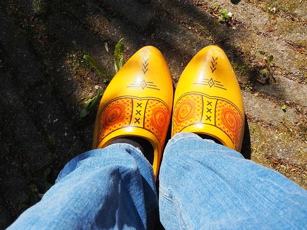 Americans see the Dutch as clog-wearers. The photo looks down at my feet: the bottoms of my jeans are visible, and the clogs are yellow with some red and black decorations. The ground is brick-paved.