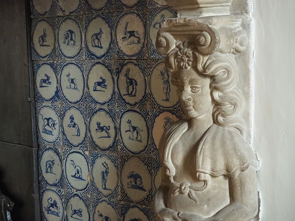 This detail shows a stone carved woman from the waist up. To her left is the back wall of the fireplace, tiled with blue and white delft tiles. 