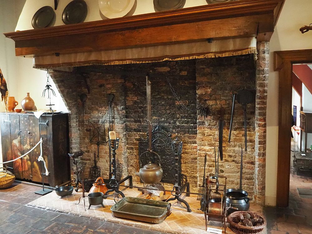 A plain brick fireplace with a wooden mantel above it. Inside it a number of kitchen utensils can be seen: copper pots and pans mostly.