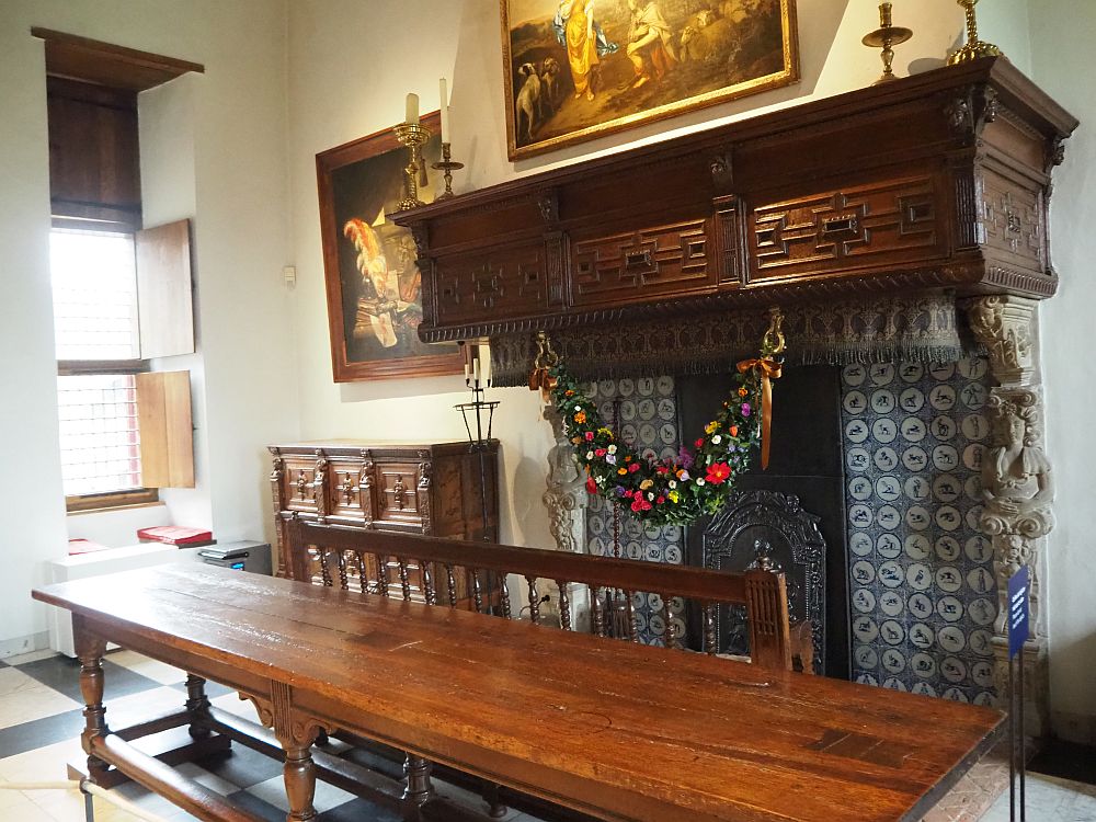 A dark wood mantelpiece over a fireplace lined with Delft tile. In front of the fireplace is a long wooden table. 