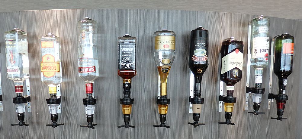 A row of 9 liquor bottles (Smirnoff, Jack Daniels, Bailey's etc.) hang upside down on a wall. Travel Risks Not Worth Taking.