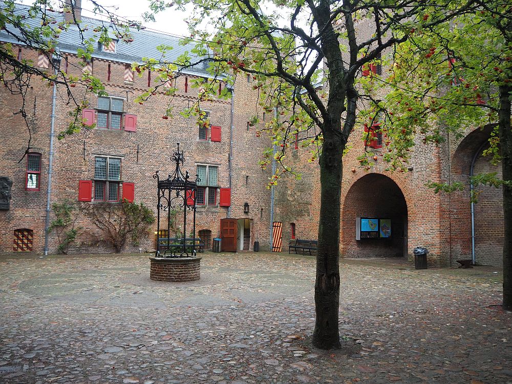 The wall beyond the courtyard is red-brick and studded with windows. A tree and a round well. An arched gateway on the right.