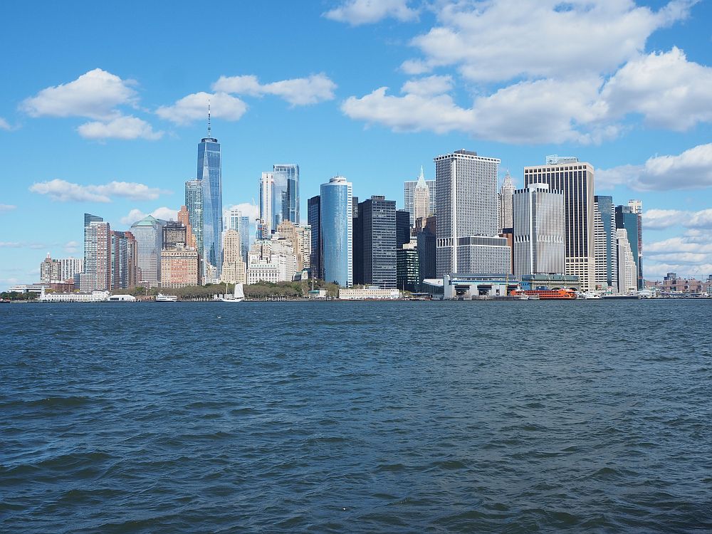 Seen across an expanse of water, the skyscrapers cluster on the horizon against a blue sky with fluffy clouds.