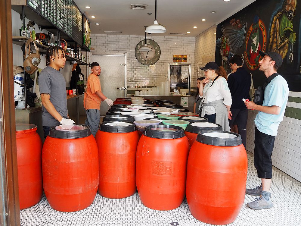 Looking into the shop, two shop workers are on the left of the room, with a row of shelving behind them. Down the center is three long rows of large barrels (thigh-high, I'd guess): bright red with a black rim at the top. On the right, several customers, and behind them on the right, a wall painted with a colorful mural, though it's hard to see what is on the mural. 