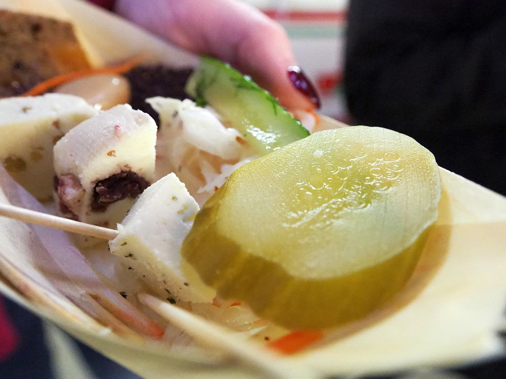 The plate full of Latvian food holds a slice of pickle and some small cubes of cheese with dark bits in them. 