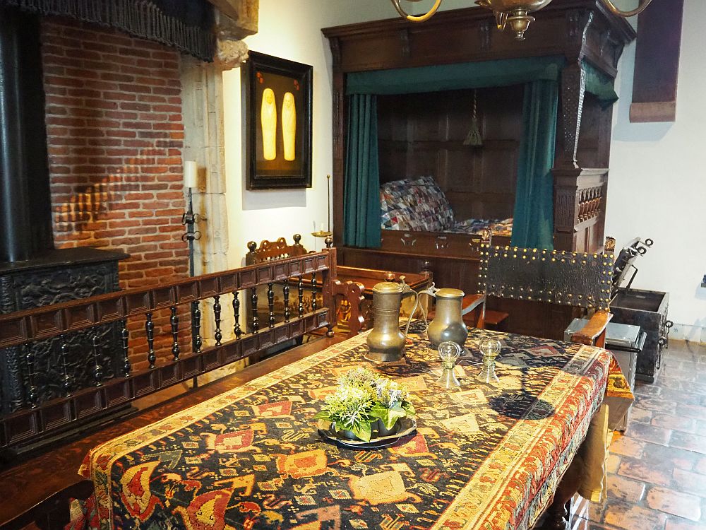 In the foreground, a table covered by a colorful rug. On the left only a corner of the large fireplace is visible. In the background is a canopied box bed.