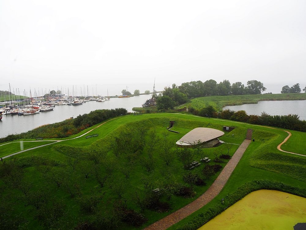 In the foreground is a grassy area with squared higher areas. Beyond that on the left is a river, and cluster of sailboats on the far side. 